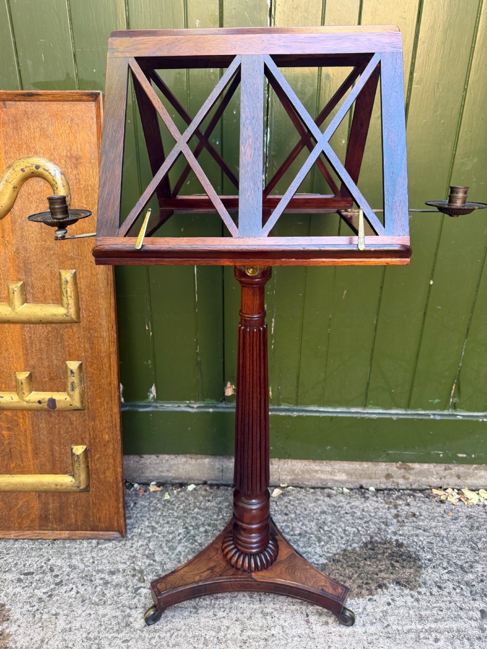 An early 19th-century Regency rosewood duet music stand featuring an adjustable double-sided frame with cross-latticed supports, twin brass candle sconces, a fluted column, and a tripartite base on castors.