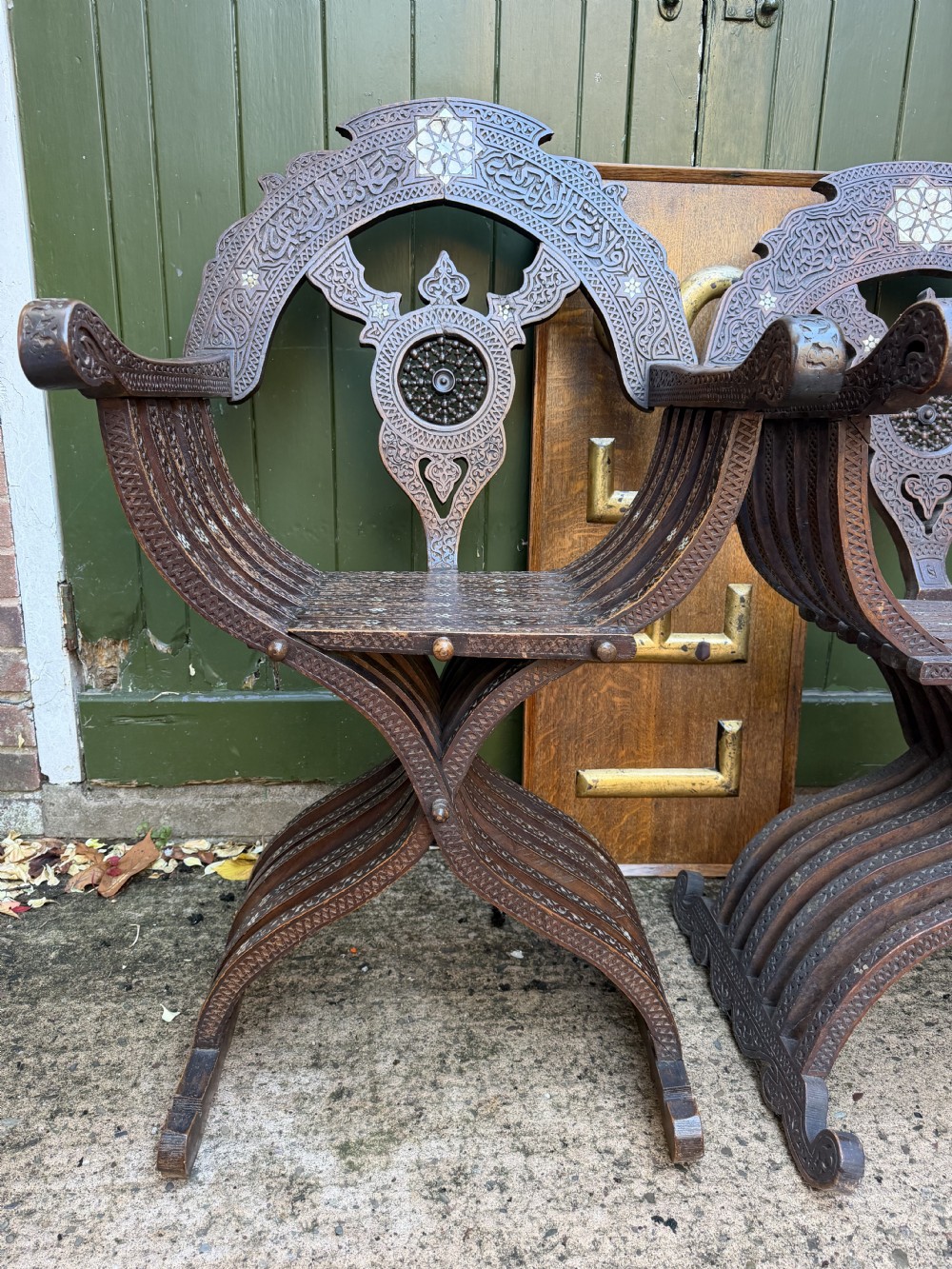 A pair of late nineteenth-century Syrian armchairs crafted from dark carved wood with mother-of-pearl inlay, Arabic script panels, and curule x-frames. View X