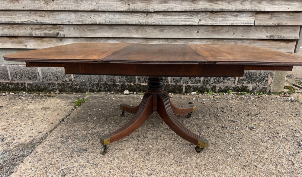 An early nineteenth-century mahogany extending dining table featuring a central reeded pedestal base with brass castors and an extra extension leaf standing behind. View XI