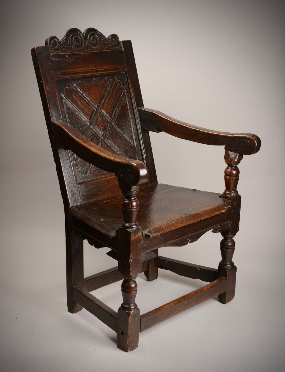 An early seventeenth-century dark oak wainscot armchair with a carved geometric panel back, turned arm supports, and a solid wooden seat.