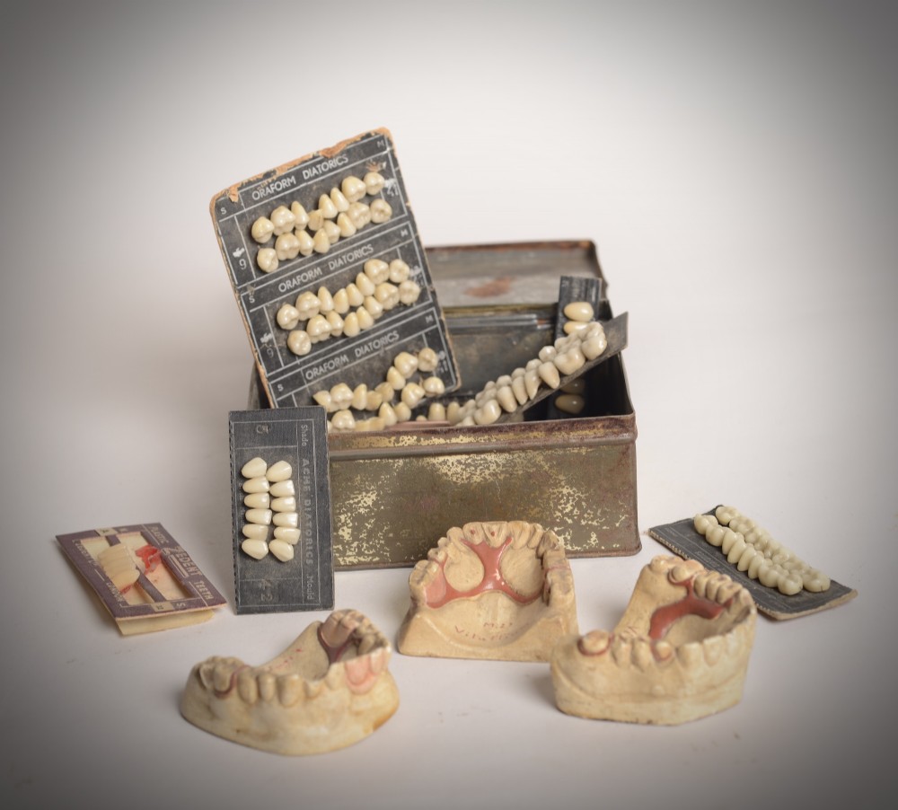 A collection of vintage dental samples featuring sets of porcelain teeth on printed cards, plaster jaw moulds, and an aged metal storage box.