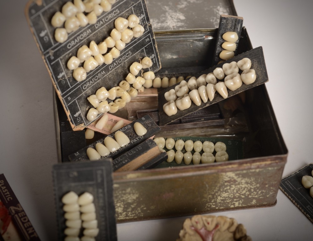 A collection of vintage dental samples featuring sets of porcelain teeth on printed cards, plaster jaw moulds, and an aged metal storage box. View III