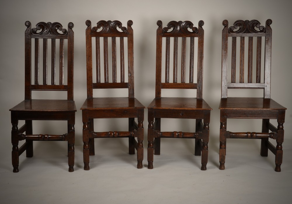 A set of four late seventeenth-century dark oak dining chairs featuring carved scroll top crestings, vertical back slats, solid panel seats, and turned stretchers.