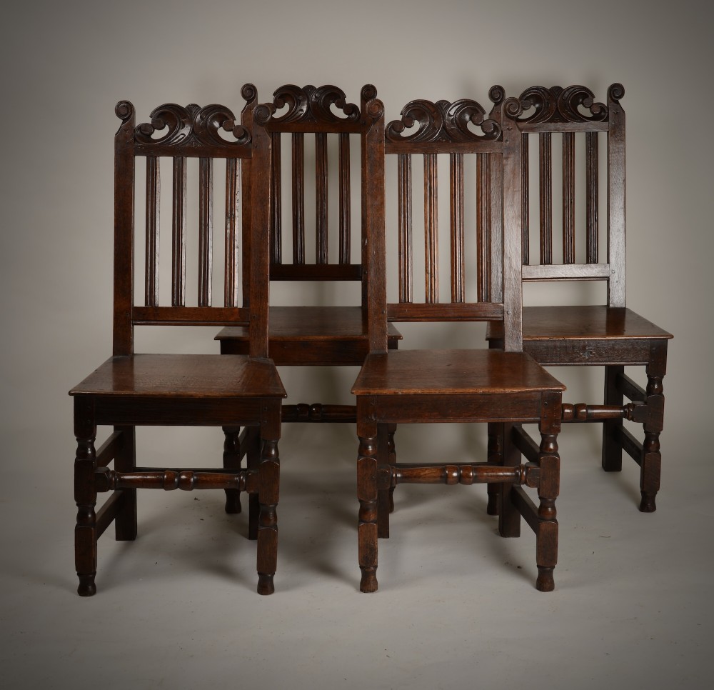 A set of four late seventeenth-century dark oak dining chairs featuring carved scroll top crestings, vertical back slats, solid panel seats, and turned stretchers. View V