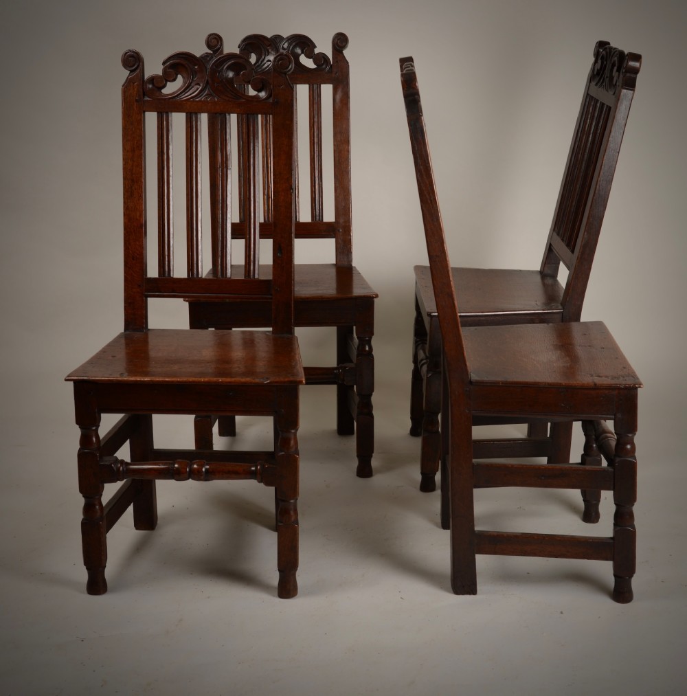 A set of four late seventeenth-century dark oak dining chairs featuring carved scroll top crestings, vertical back slats, solid panel seats, and turned stretchers. View VI