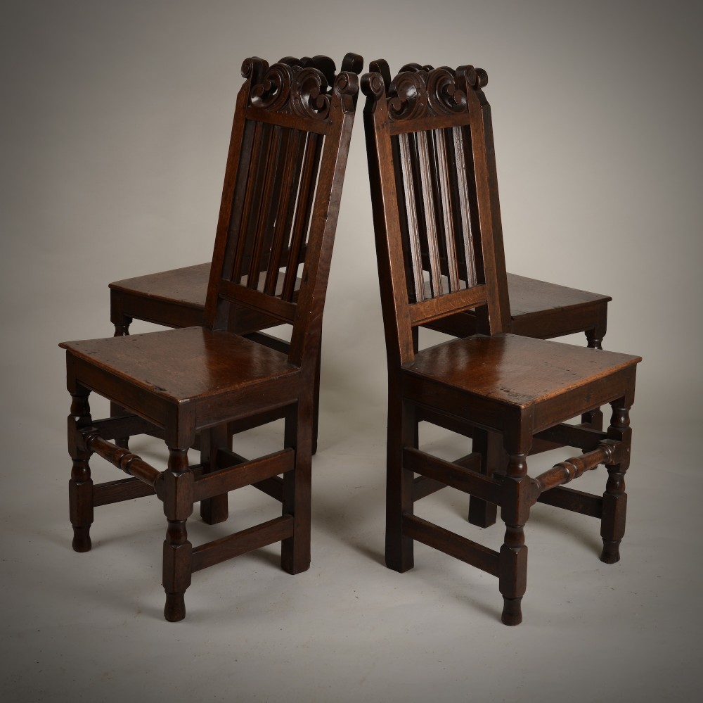 A set of four late seventeenth-century dark oak dining chairs featuring carved scroll top crestings, vertical back slats, solid panel seats, and turned stretchers. View VII