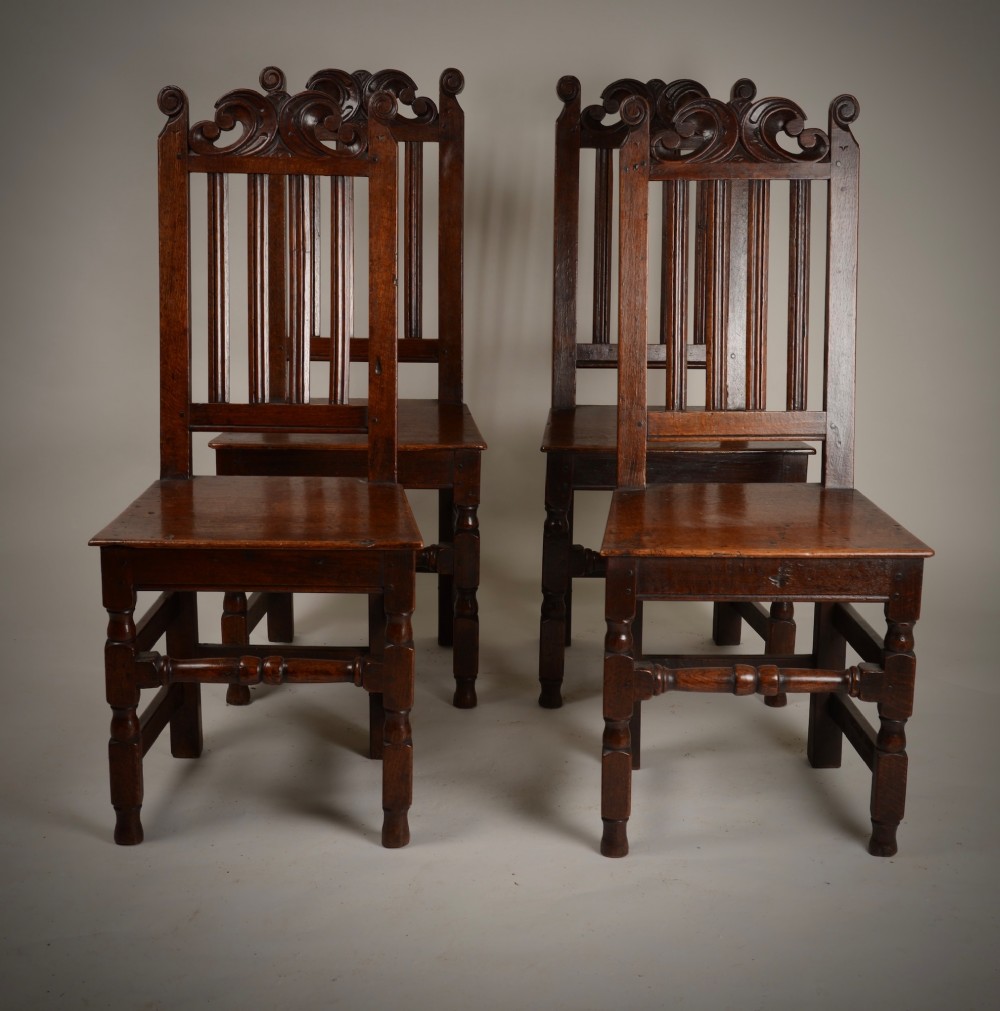 A set of four late seventeenth-century dark oak dining chairs featuring carved scroll top crestings, vertical back slats, solid panel seats, and turned stretchers. View VIII