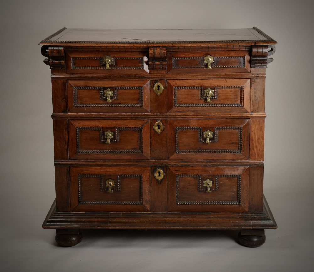 An antique warm brown walnut chest of drawers featuring geometric moulded drawer fronts, brass drop handles, and turned bun feet.