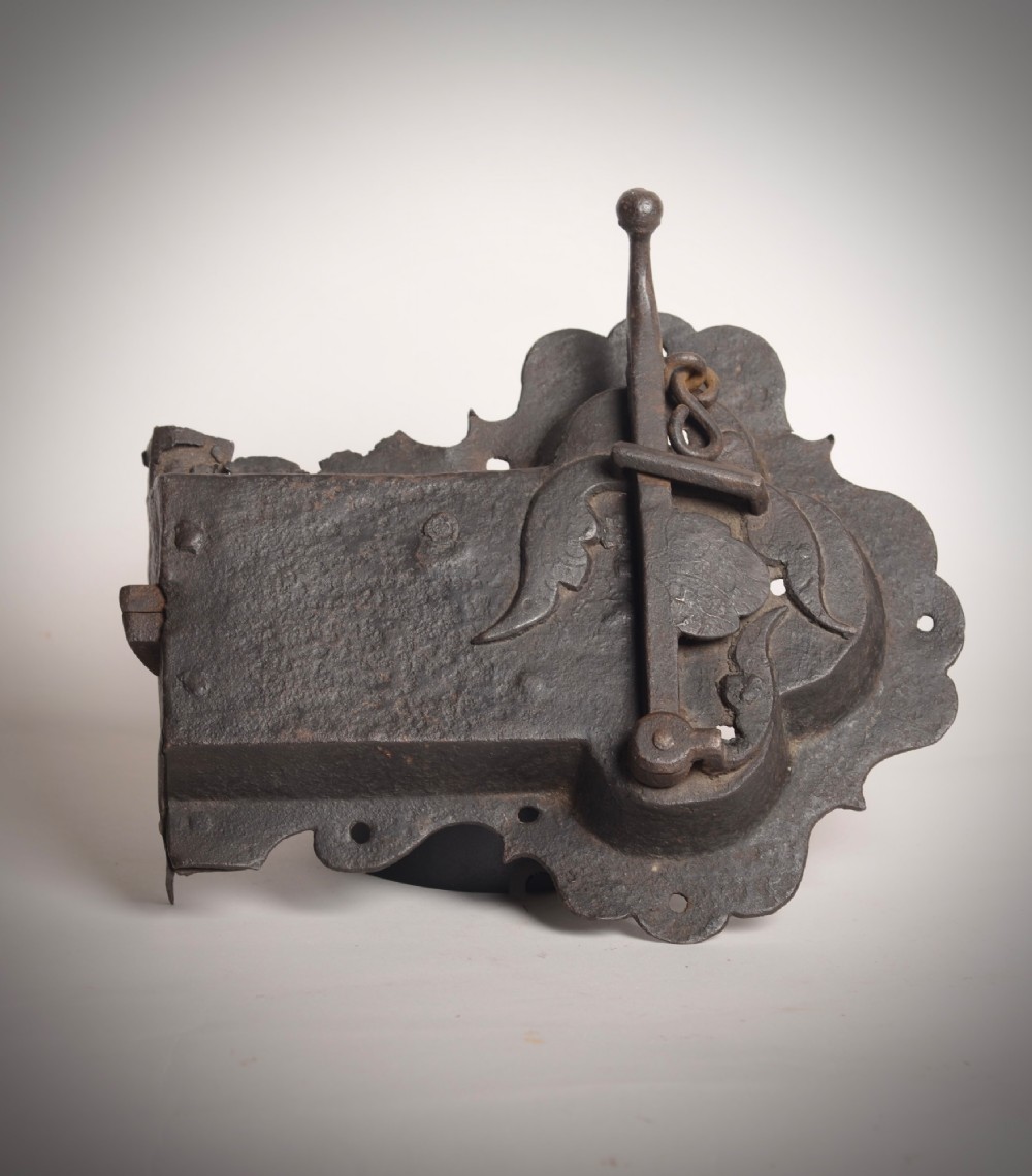 A 17th-century hand-forged wrought iron rim lock featuring a scalloped decorative backplate, a surface-mounted lever latch, and aged dark metal patina.