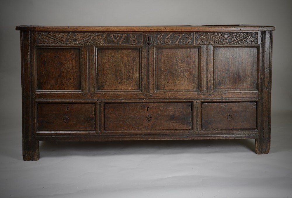 A seventeenth-century dark oak mule chest featuring four carved front panels, three lower drawers, and a carved top frieze with the initials WB and date 1679.