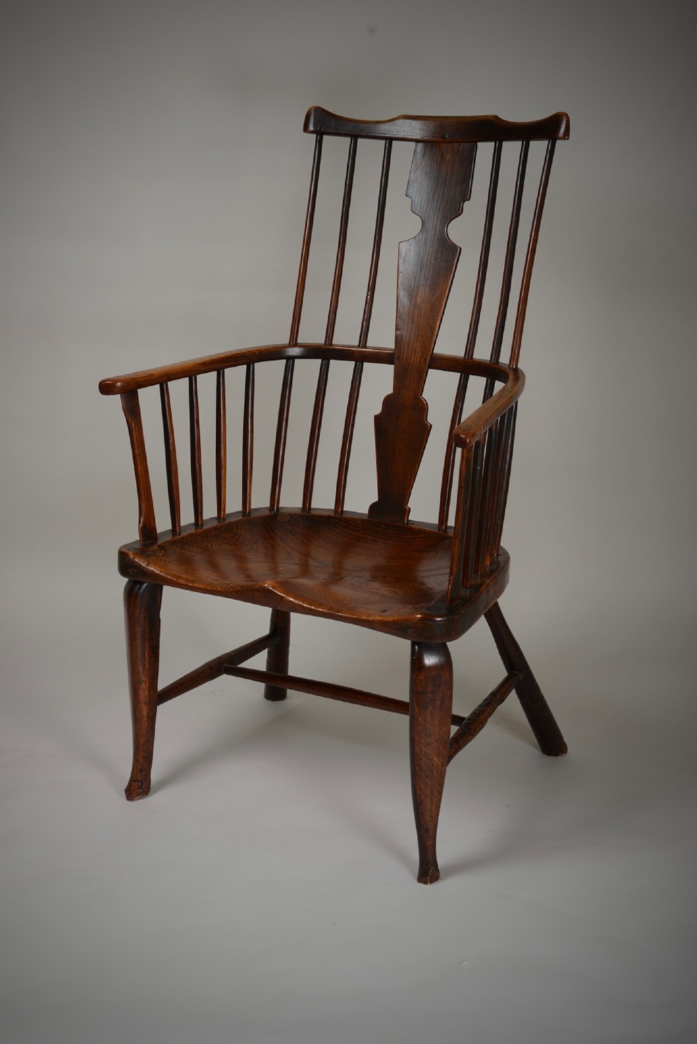 A dark brown antique wooden comb-back Windsor armchair featuring a shaped central splat, slim spindles, a contoured saddle seat, and turned legs.