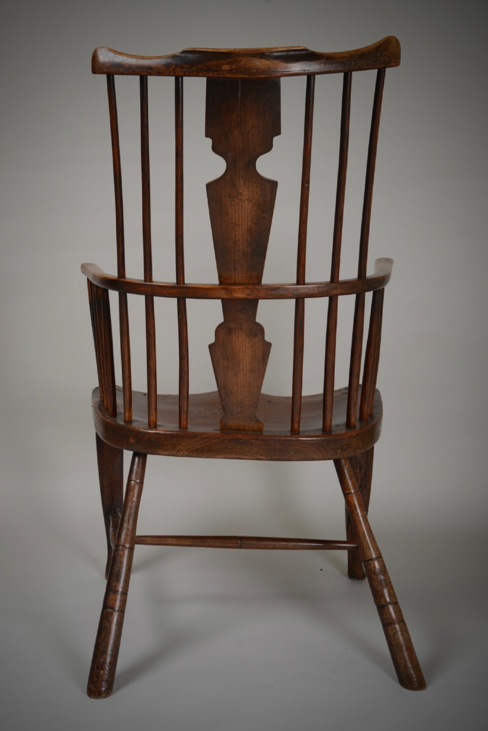 A dark brown antique wooden comb-back Windsor armchair featuring a shaped central splat, slim spindles, a contoured saddle seat, and turned legs. View X