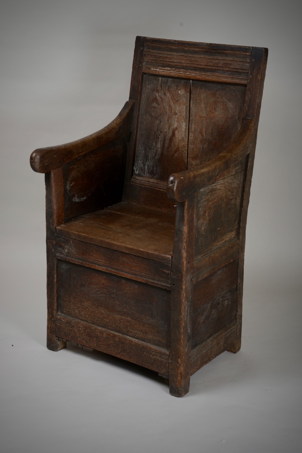 A dark oak late Elizabethan box chair featuring a high panelled back, curved armrests, and an enclosed wooden base.