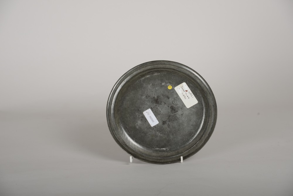 A circular seventeenth-century pewter dish featuring a raised reeded rim, shallow well, dark patinated surface, and stamped initial markings. View II