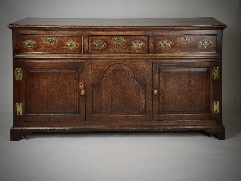 An 18th-century dark oak dresser base featuring three drawers over two cupboard doors flanking a central arched fielded panel with brass hardware.