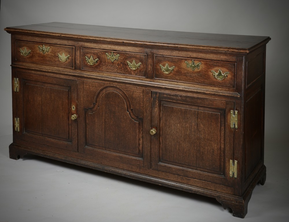 An 18th-century dark oak dresser base featuring three drawers over two cupboard doors flanking a central arched fielded panel with brass hardware. View V