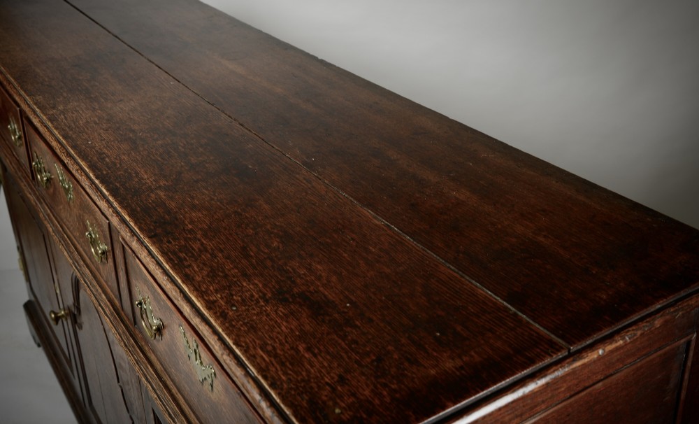 An 18th-century dark oak dresser base featuring three drawers over two cupboard doors flanking a central arched fielded panel with brass hardware. View VI