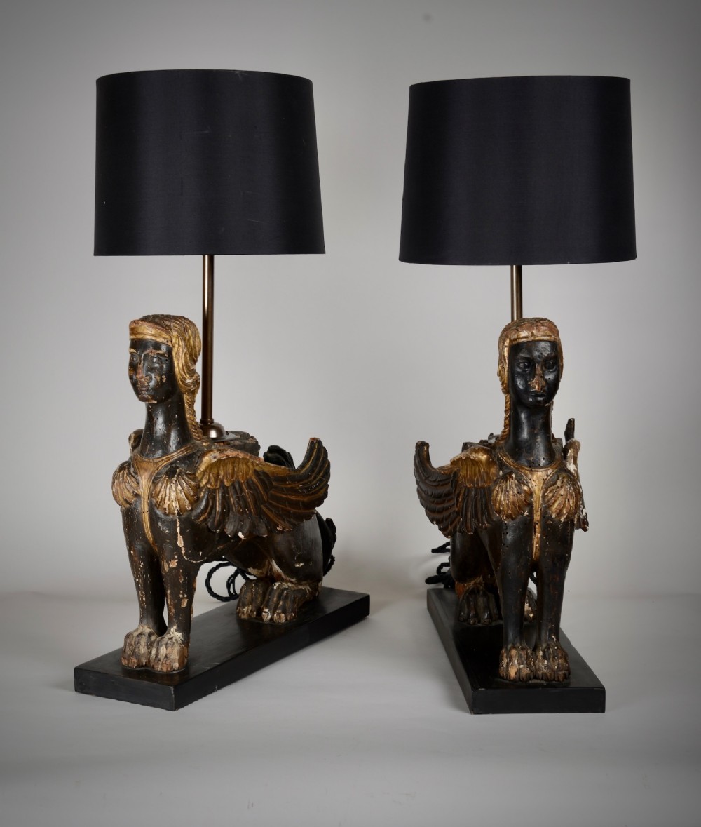 A pair of 18th-century ebonised and parcel-gilt carved wooden sphinx sculptures mounted on rectangular plinths and converted into table lamps with black shades.