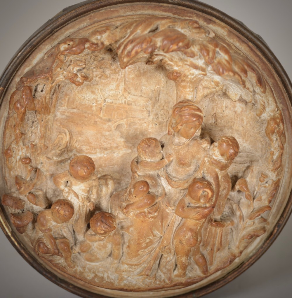 A round beige plaster relief capturing figures carved in high relief, secured inside a circular metal frame with a hanging loop at the top. View II