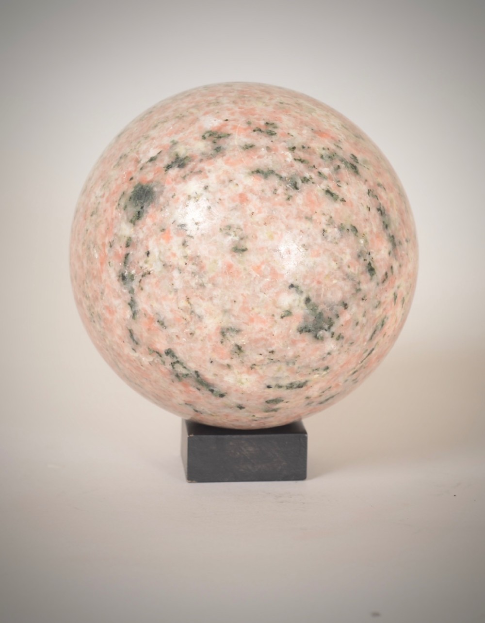 A polished pink granite sphere with grey speckled inclusions rests on a small dark square base. View II