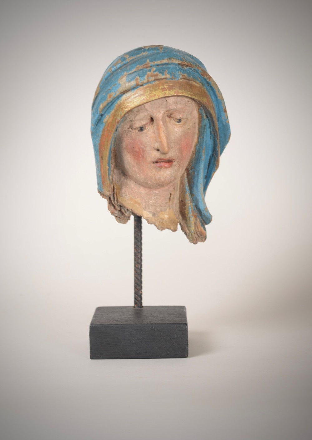 A 17th-century carved wooden head fragment of the Virgin Mary featuring polychrome blue and gold headwear, mounted on a slim black metal rod and square base.