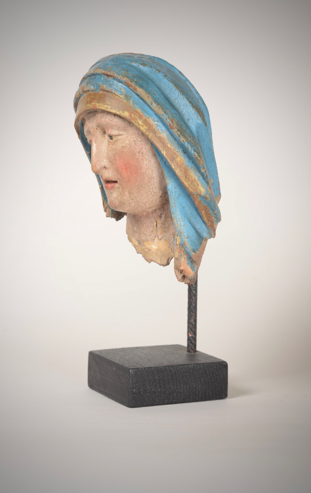 A 17th-century carved wooden head fragment of the Virgin Mary featuring polychrome blue and gold headwear, mounted on a slim black metal rod and square base. View II