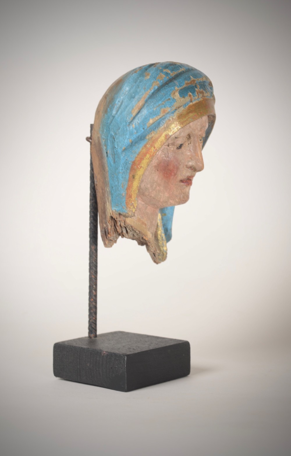 A 17th-century carved wooden head fragment of the Virgin Mary featuring polychrome blue and gold headwear, mounted on a slim black metal rod and square base. View IV