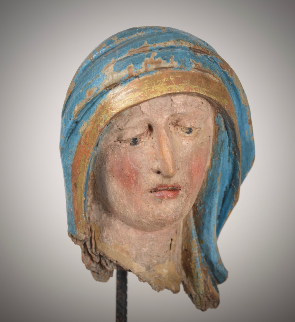 A 17th-century carved wooden head fragment of the Virgin Mary featuring polychrome blue and gold headwear, mounted on a slim black metal rod and square base. View V