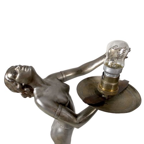 An Art Deco spelter table lamp featuring a silvered female figure holding up an amber glass globe shade on a circular onyx base. View X