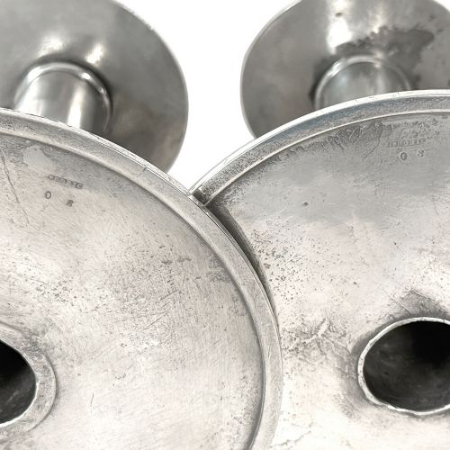 A pair of silver-tone pewter candlesticks featuring tapered cylindrical stems, flat wide sconces, and broad circular bases decorated with relief stylised plant motifs. View VI