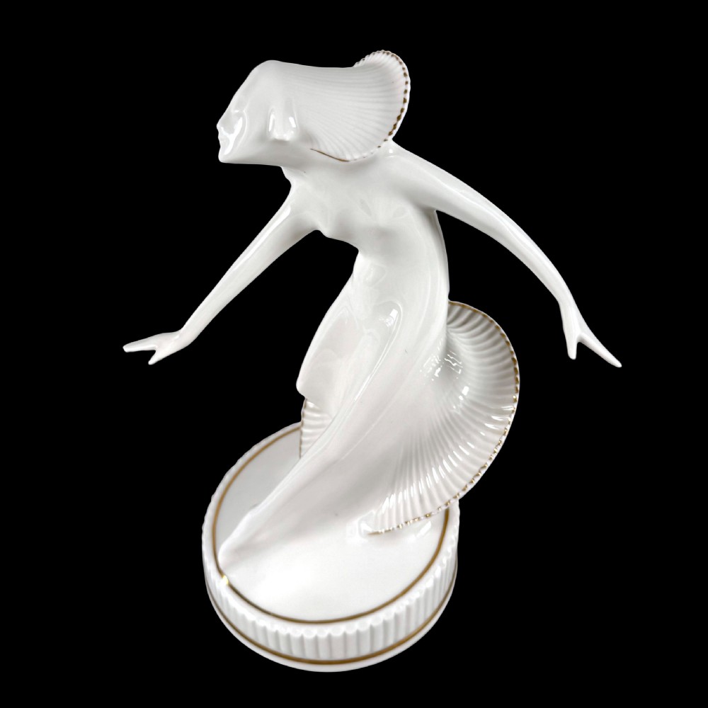 A white glazed porcelain Art Deco figurine depicting a female dancer in dynamic motion with pleated gold-trimmed drapery on a circular base.
