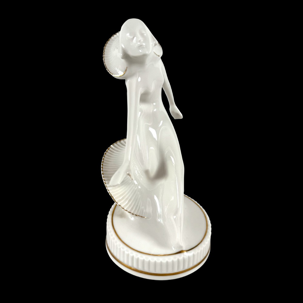 A white glazed porcelain Art Deco figurine depicting a female dancer in dynamic motion with pleated gold-trimmed drapery on a circular base. View IV