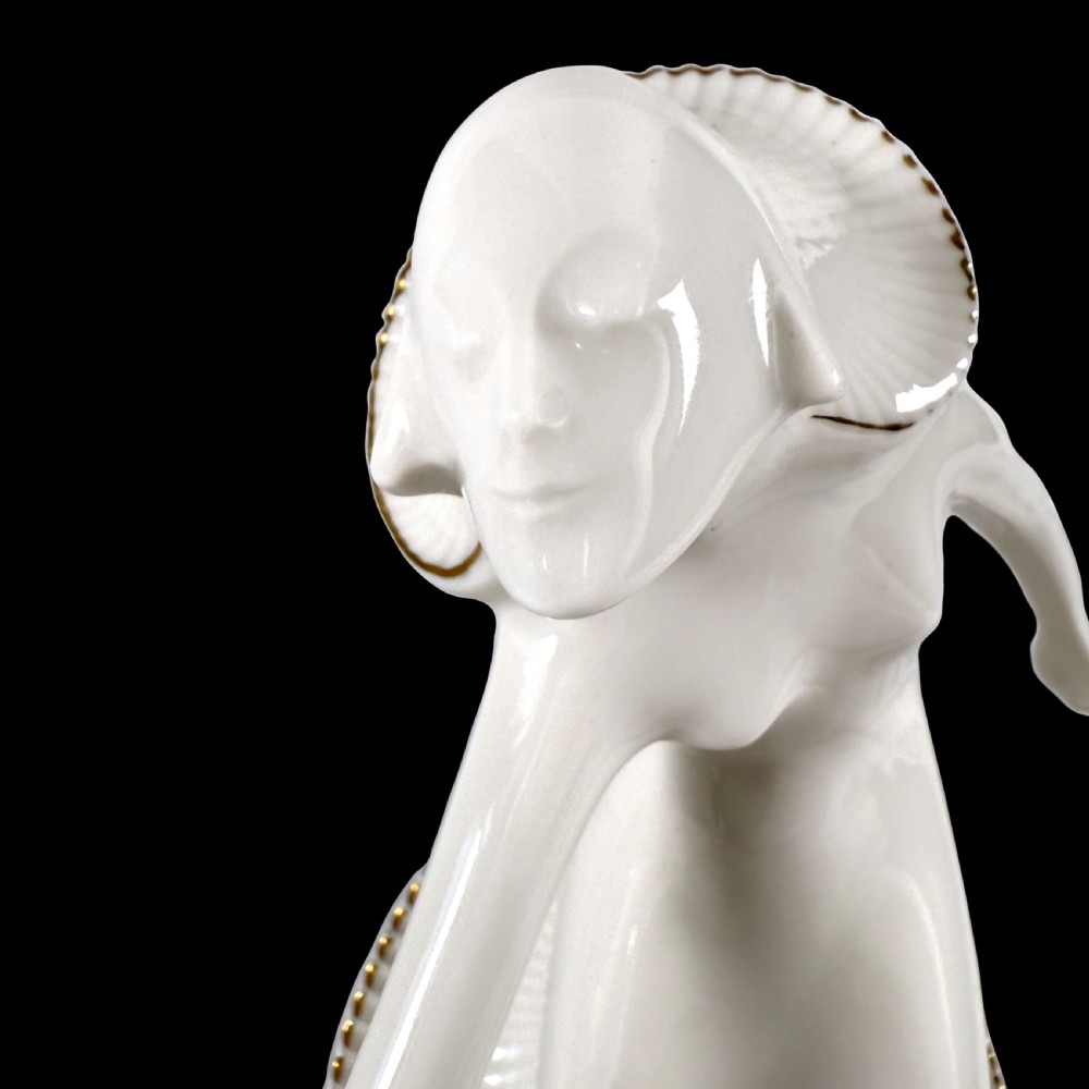 A white glazed porcelain Art Deco figurine depicting a female dancer in dynamic motion with pleated gold-trimmed drapery on a circular base. View VII