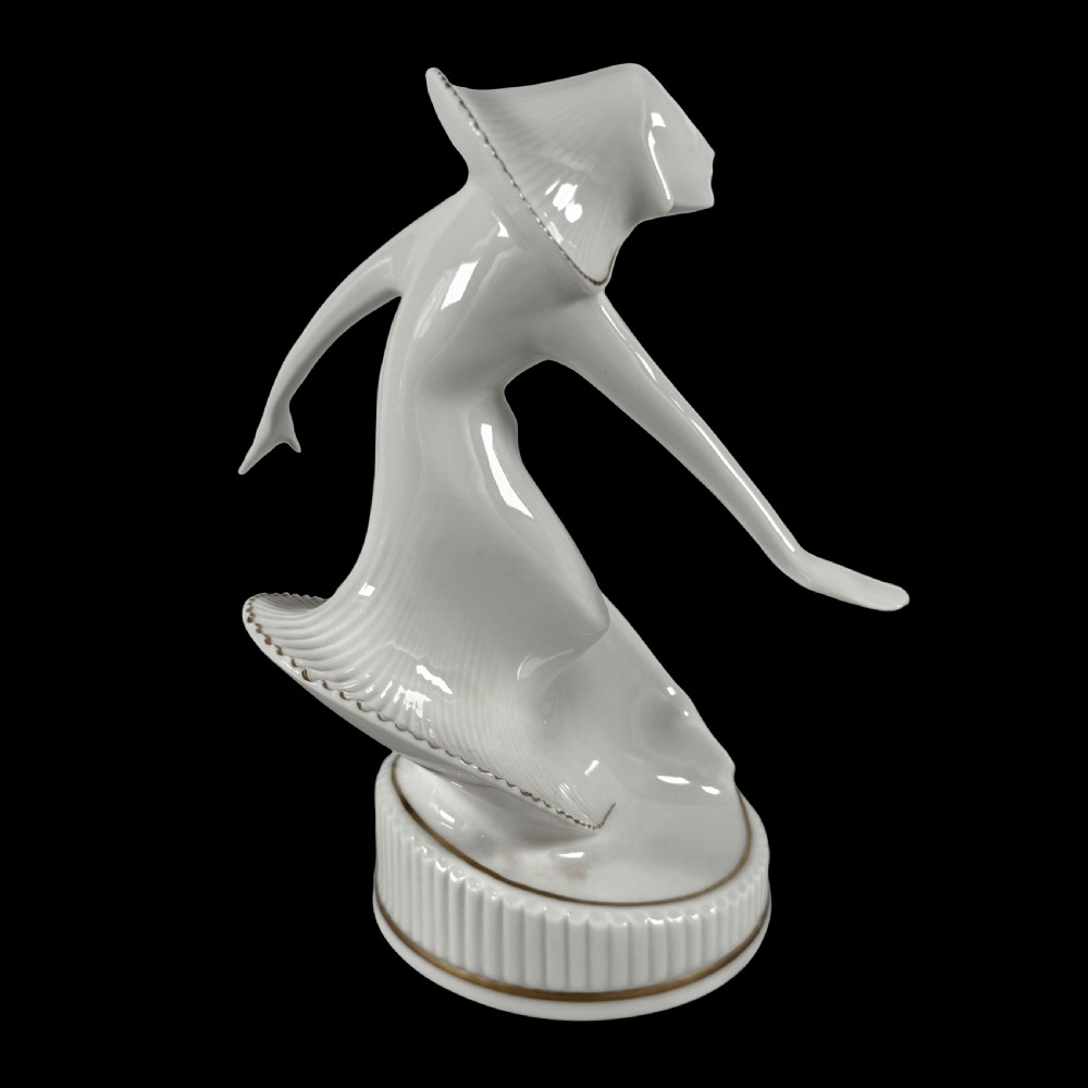 A white glazed porcelain Art Deco figurine depicting a female dancer in dynamic motion with pleated gold-trimmed drapery on a circular base. View IX