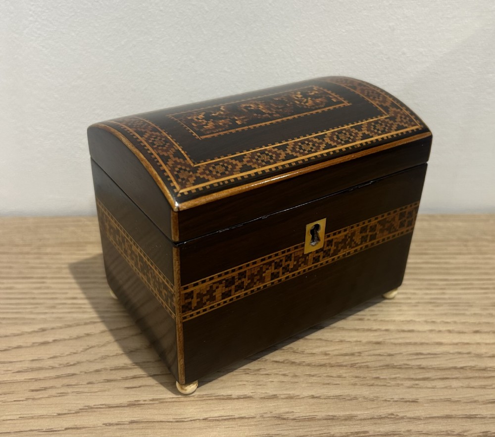 A Victorian rosewood tea caddy featuring a domed lid, geometric mosaic wood inlay, and a brass keyhole escutcheon. View VI