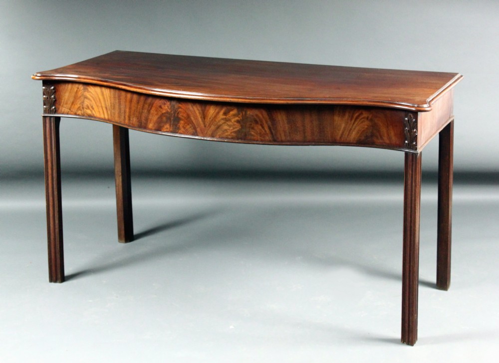 An antique Georgian Chippendale mahogany side table featuring a serpentine-curved frieze with flame mahogany veneers and straight reeded legs.