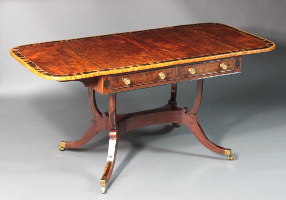 A matching three-piece Regency period furniture set featuring a central sofa table with drawers flanked by a pair of D-shaped folding card tables, all crafted with figured wood and crossbanded inlay borders on curved legs with brass castors. View V