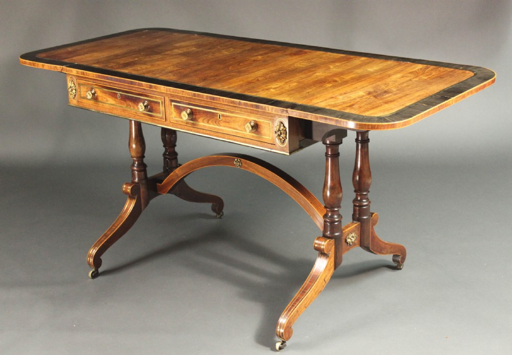 A Regency period kingwood sofa table with brass inlay, two drawers, drop-leaf sides, turned legs, an arched stretcher, and brass castors.