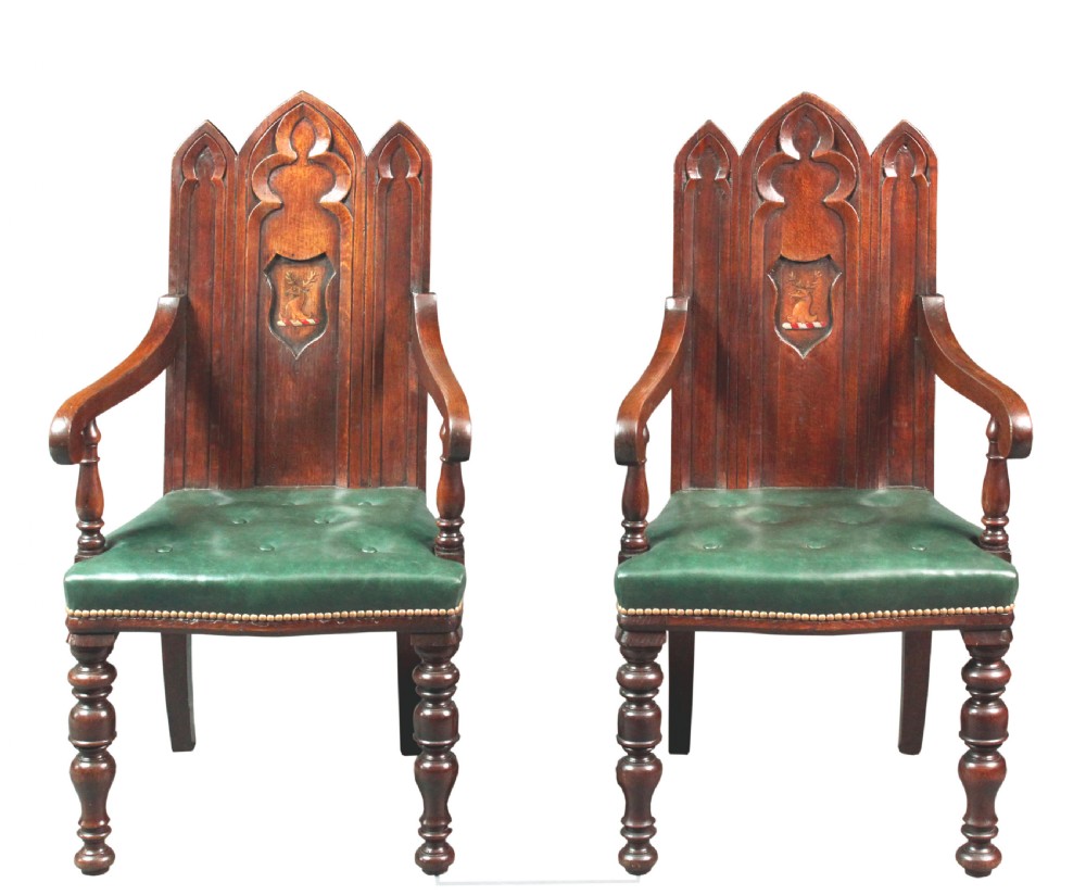 A pair of carved dark oak Gothic Revival hall armchairs featuring pointed arch backrests with painted armorial crests and green leather seats.
