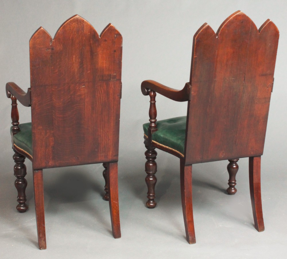 A pair of carved dark oak Gothic Revival hall armchairs featuring pointed arch backrests with painted armorial crests and green leather seats. View VII