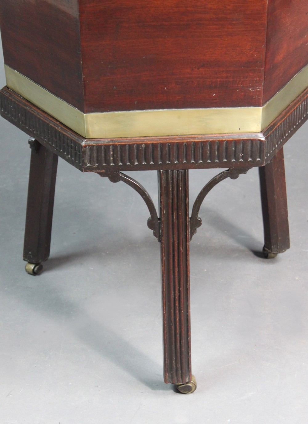 A hexagonal mahogany wine cooler featuring polished brass bands, side handles, and a fitted stand with fluted legs and brass castors. View II