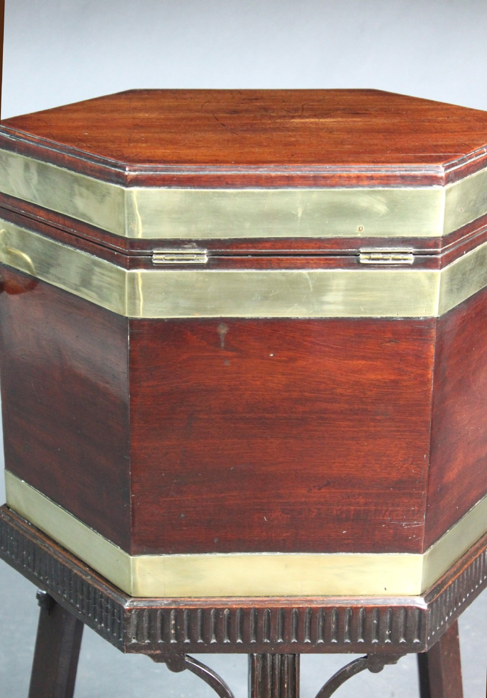 A hexagonal mahogany wine cooler featuring polished brass bands, side handles, and a fitted stand with fluted legs and brass castors. View IV