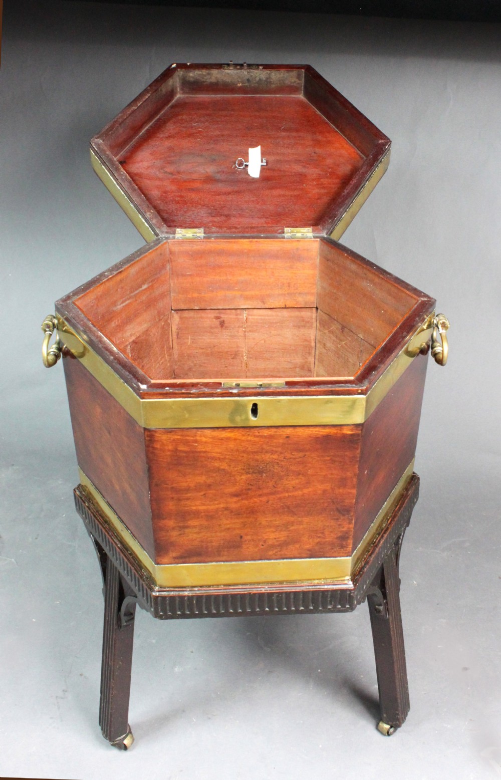 A hexagonal mahogany wine cooler featuring polished brass bands, side handles, and a fitted stand with fluted legs and brass castors. View VII