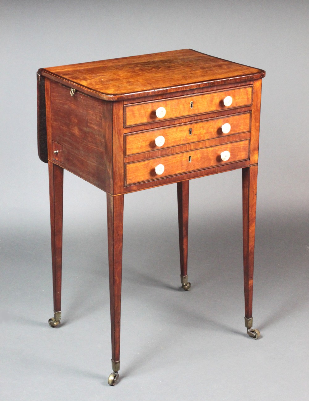 A George III satinwood sewing table featuring three drawers with bone knobs, hinged drop leaves, and square tapered legs on brass castors.