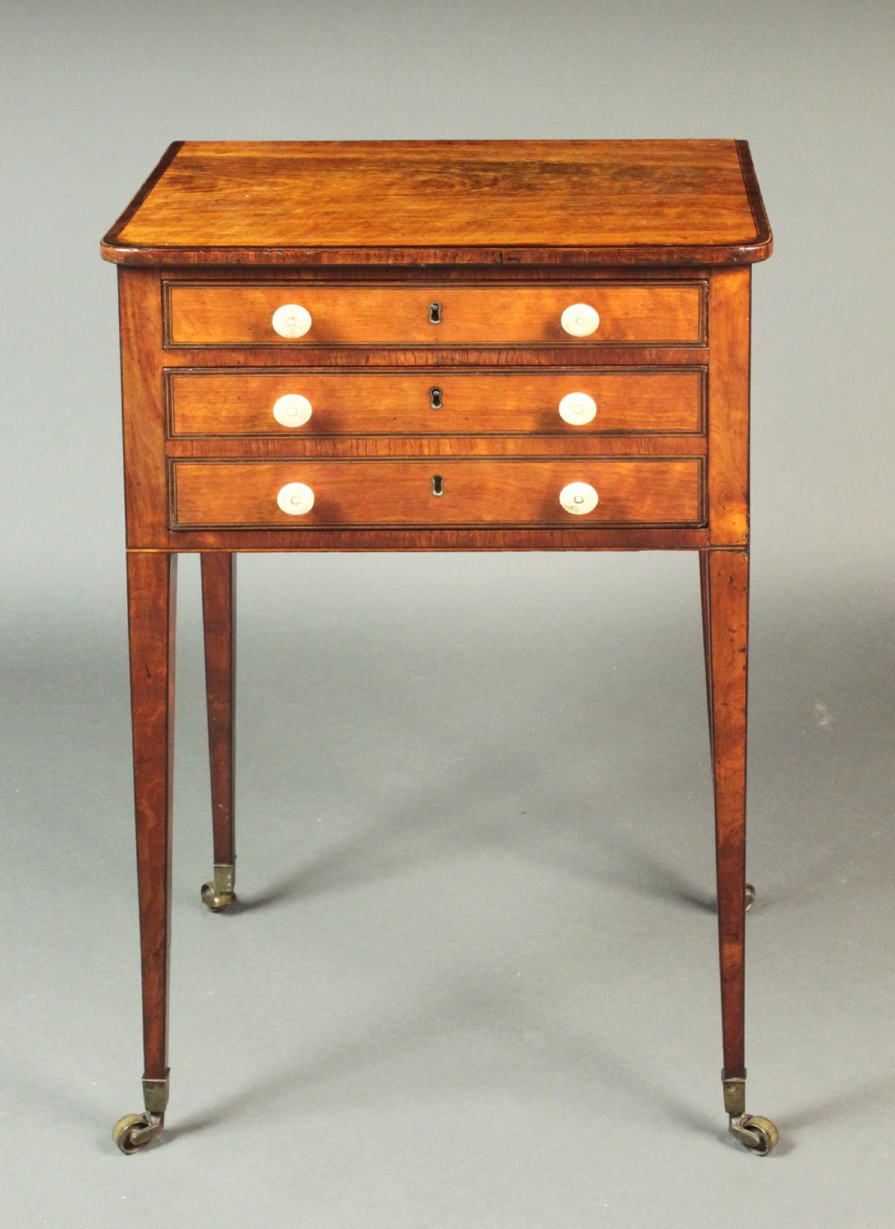 A George III satinwood sewing table featuring three drawers with bone knobs, hinged drop leaves, and square tapered legs on brass castors. View II