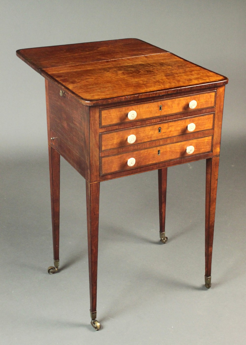 A George III satinwood sewing table featuring three drawers with bone knobs, hinged drop leaves, and square tapered legs on brass castors. View III