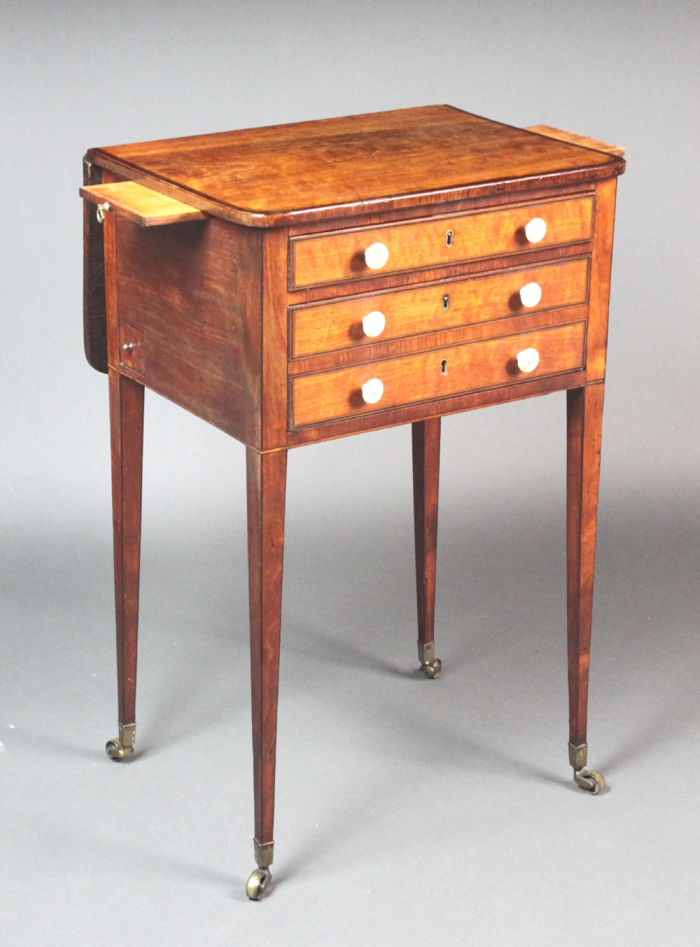 A George III satinwood sewing table featuring three drawers with bone knobs, hinged drop leaves, and square tapered legs on brass castors. View V