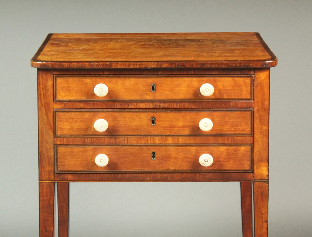A George III satinwood sewing table featuring three drawers with bone knobs, hinged drop leaves, and square tapered legs on brass castors. View VI