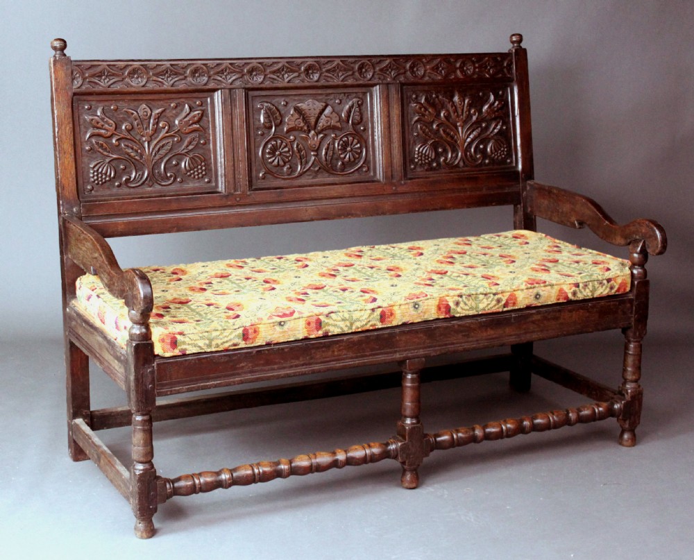 A late seventeenth-century dark carved oak settle featuring three carved back panels with floral motifs, turned legs, stretchers, and a floral patterned seat cushion.