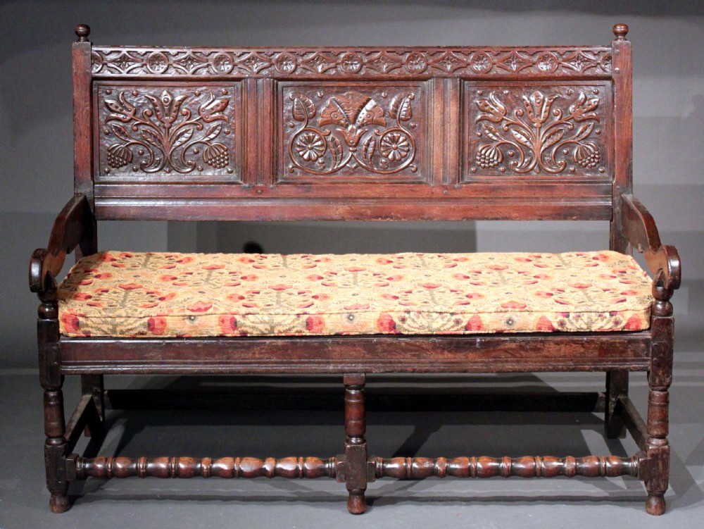 A late seventeenth-century dark carved oak settle featuring three carved back panels with floral motifs, turned legs, stretchers, and a floral patterned seat cushion. View II